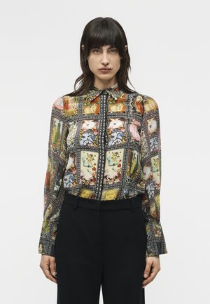 Nő, aki színes, mintás, hosszú ujjú, gombos blúzt visel, amelyen tarot-kártya minták találhatók, és fekete nadrágot, egyszínű háttér előtt.