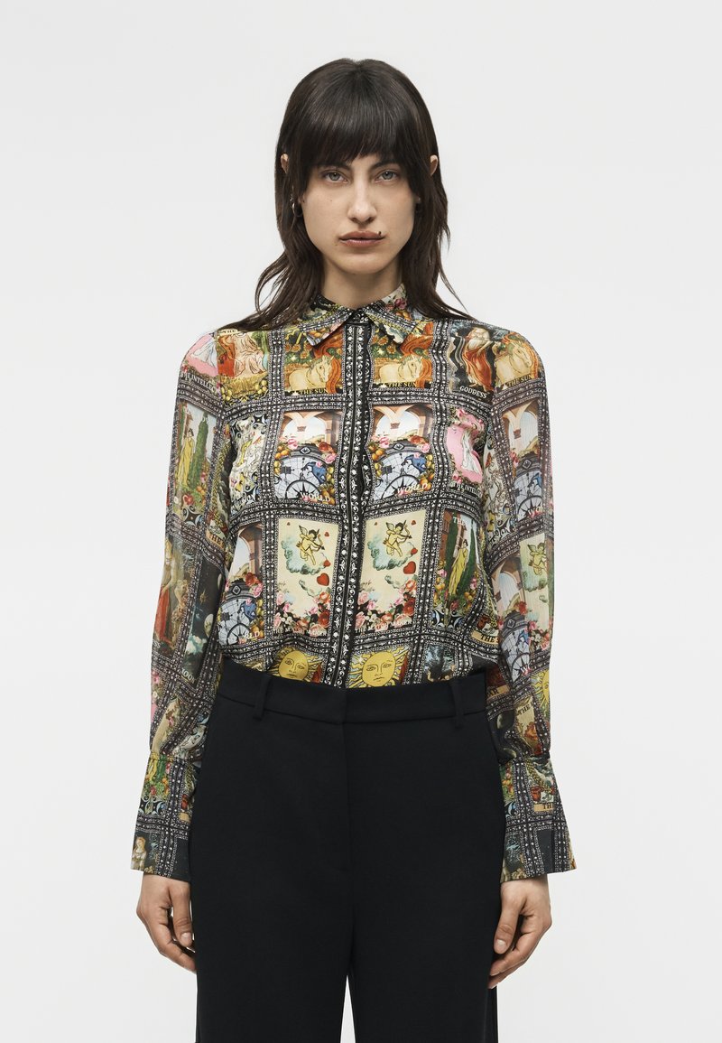 Γυναίκα που φοράει πολύχρωμη, μακριάζ υφασμάτινη μπλούζα με κουμπιά και μοτίβα ταρώ καρτών, καθώς και μαύρο παντελόνι, μπροστά σε ένα μονόχρωμο φόντο.