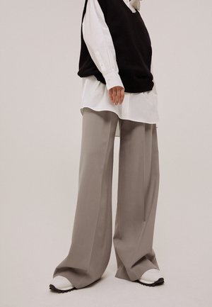 Persona que lleva pantalones anchos beige, zapatillas blancas, una camisa blanca larga y un chaleco de punto sin mangas negro sobre un fondo liso.