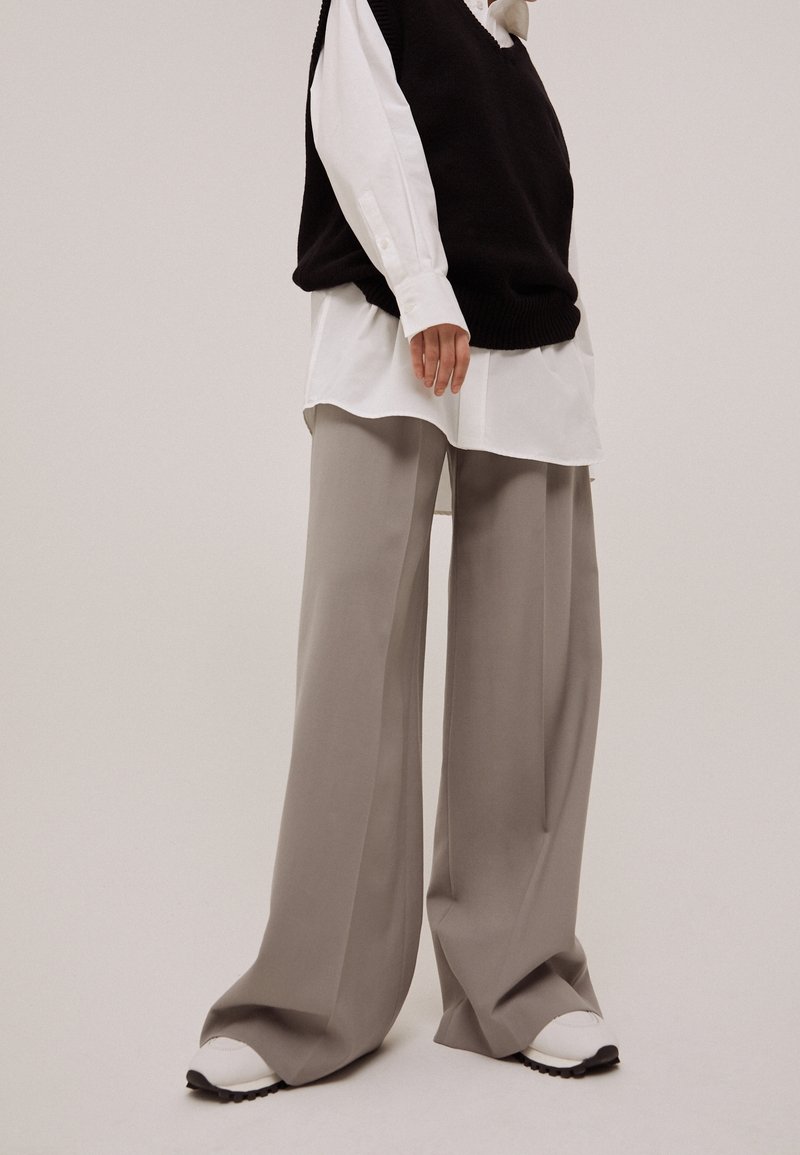 Personne portant un pantalon large beige, des baskets blanches, une longue chemise blanche et un gilet sans manches noir sur un fond uni.