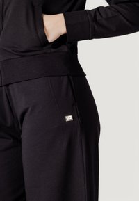 Completo sportivo nero con una parte superiore con zip e pantaloni affusolati. I dettagli notevoli includono un piccolo logo metallico sui pantaloni e il design della tasca.