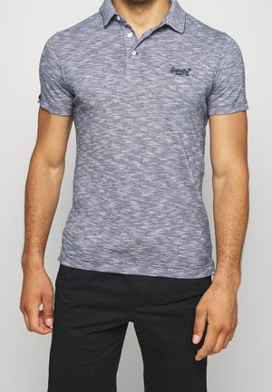 Mann mit kurzärmeligem, blau-grau gemustertem Poloshirt mit Kragen und schwarzer Hose, steht vor einem schlichten, hellen Hintergrund.
