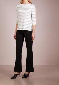 Frau in einem weißen, gestreiften Langarmoberteil, schwarzen, figurbetonten Hosen und schwarzen, spitzen Absatzmules, die auf einem beigen Boden steht.