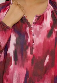 Leichtes rotes und lila gemustertes Blusenshirt mit V-Ausschnitt, gerafften Ärmeln und dekorativen Knöpfen entlang der Knopfleiste. Strukturierter Stoff.