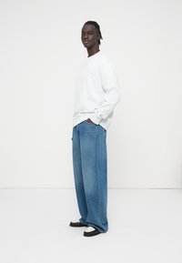 Ανοιχτό γκρι φούτερ με μακριά μανίκια και ριμπ μανσέτες, σε συνδυασμό με φαρδιά, μεσαίου μπλε τζιν παντελόνια. Μαύρα και άσπρα παπούτσια με χοντρές σόλες.