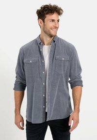 Graues Cordhemd mit Knopfleiste, zwei Brusttaschen und hochgekrempelten Ärmeln. Weicher Stoff und lockere Passform. Über einem weißen T-Shirt getragen.