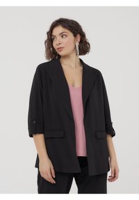 Blazer nero con un design strutturato, caratterizzato da ampie rever, maniche arrotolate e tasche frontali, indossato sopra una maglietta rosa.