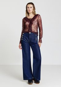 Blusa di pizzo color borgogna con dettagli arricciati e fiocco, abbinata a jeans di denim a vita alta a gamba larga con bottoni dorati.
