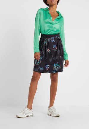 Vrouw die een glanzende groene blouse met lange mouwen draagt, een zwarte gebloemde knielange rok, en witte chunky sneakers, staand op een effen witte achtergrond.