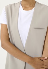 Beige ärmellose Weste mit glatter Textur, ausgestattet mit einer schmalen Brusttasche und getragen über einem weißen T-Shirt.