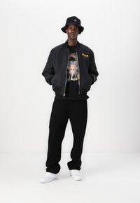 Giubbotto bomber nero con logo giallo "Kodak", t-shirt grafica, pantaloni neri e sneakers bianche. Un cappello a secchiello completa il look.