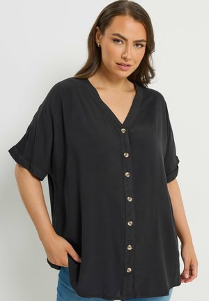 Kvinde iført en løstsiddende sort skjorte med knapper, korte op-rullede ærmer og V-hals, poserer med den ene hånd i lommen.