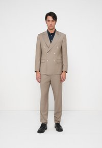 Ανδρας που φοράει μπεζ σακάκι διπλής όψης με μαύρο πουκάμισο και μαύρα επίσημα παπούτσια, σε λευκό φόντο.