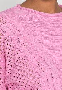 Pink knitted sweater featuring a textured cable design and open knit pattern on one side. Smooth finish with a round neckline.