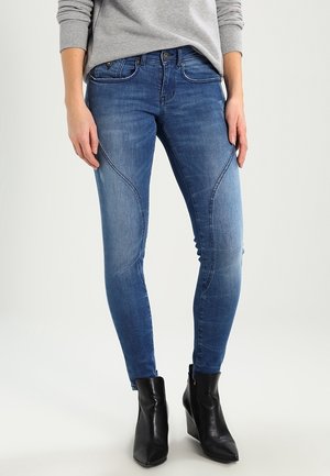 Frau mit blauen Skinny-Jeans mit geschwungenen Nähten und schwarzen Knöchelstiefeln, die vor einem hellen Hintergrund steht.