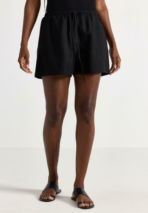 Person iført sorte shorts med snøre og sorte slip-on sandaler, stående mod en ensfarvet lys baggrund.