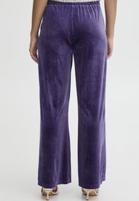 Fioletowe welurowe spodnie o szerokich nogawkach z elastycznym pasem, gładką fakturą i lekkim połyskiem, zestawione z obcasami w neutralnych kolorach.