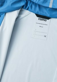 Blå jakke med en glat overflade og hvidt indre lag. Indeholder et mærke, der viser bæredygtigt design, størrelse 104 cm, og klip linjer.