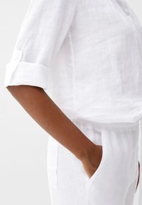 Weiße Leinenbluse mit aufgerollten Ärmeln, mit lässiger Passform und Knopfdetails, kombiniert mit weißen Leinenhosen, die eine Hand in der Tasche hat.