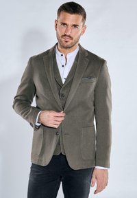 Olivgrüner Blazer aus einem strukturierten Stoff, mit Reverskragen, zwei vorderen Taschen und einer einreihigen Knopfschließe, über einem weißen Hemd getragen.