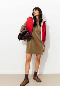 Gilet matelassé rouge, robe chemise en velours côtelé olive, chemise en flanelle à motifs, chaussettes côtelées marron et chaussures à imprimé léopard marron. Fond minimaliste.