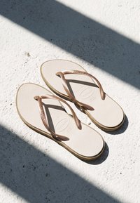 Par beige flip-flops med rosegullstropper som hviler på en hvit teksturert overflate med diagonale skygger.