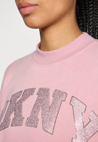 Rózsaszín pulóver magas nyakkal, nagy csillámos "DKNY" felirattal, ezüst strasszokkal, sima felülettel és laza fazonnal.