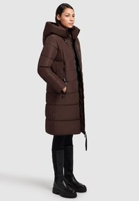 Un manteau rembourré marron avec une capuche, un design à coutures verticales, deux zips latéraux et une taille légèrement cintrée, associé à des bottes noires jusqu'aux genoux.