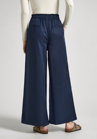 Kvinna iklädd marinblå vida byxor med elastisk midja och beige sandaler, stående mot en enfärgad ljus bakgrund.