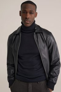 Svart läderbomberjacka med dragkedja, ståkrage, ribbade muddar, bärs över en mörk polotröja, kombinerad med grå byxor.