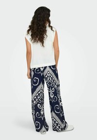 Top blanc sans manches associé à un pantalon large marine avec un motif tourbillonnant blanc et un tissu texturé. Des chaussures décontractées complètent le look.