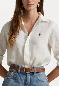 Bílá lněná košile s knoflíkovým zapínáním, malým námořnickým logem na prsou, kombinována se sepranými modrými džíny a hnědým koženým páskem se zlatou sponou.