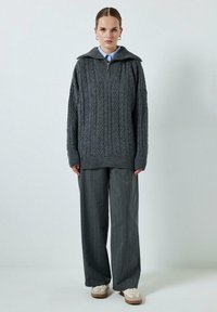 Šedý svetr s úpletem a vysokým límcem, s detailem na zip, doplněný šedými pruhovanými kalhotami s širokými nohavicemi a smetanovými teniskami.