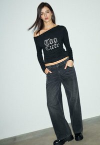 Schwarzes Langarmshirt mit Strassschriftzug "Zu süß", kombiniert mit weiten schwarzen Jeans und klobigen schwarzen Stiefeln, auf einem neutralen Hintergrund.