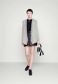 Cardigan gris clair sur un haut noir, associé à une jupe en cuir noire avec des boutons, des chaussures noires à enfiler et un sac à carreaux noir et blanc.