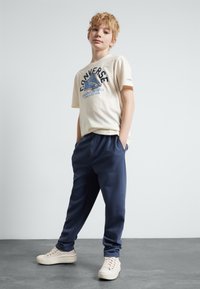 T-shirt en coton beige avec impression "Converse", pantalon élastique bleu marine et baskets beige clair. Tenue décontractée avec une coupe ample.