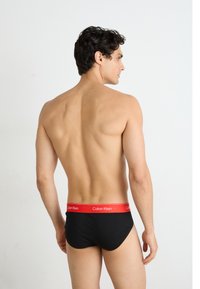 Fekete pamut alsónadrág piros gumírozott derékkal, rajta a "Calvin Klein" logóval. A modell háttal áll a kamerának.