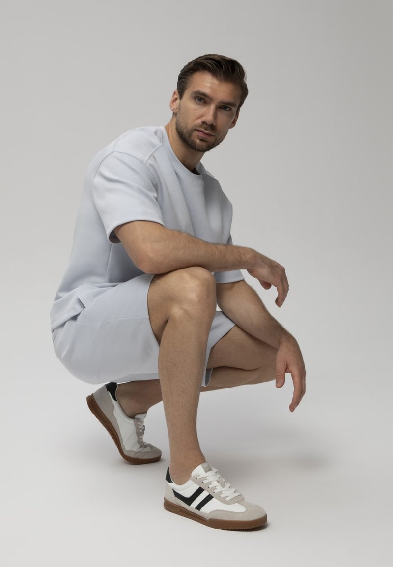 Homme portant une chemise à manches courtes gris clair, un short assorti et des baskets blanches avec des rayures noires, accroupi sur un fond clair uni.