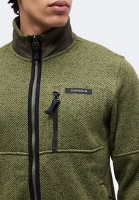 Homme portant une veste zippée texturée verte avec une fermeture éclair noire et une poche poitrine, écusson logo Icepeak sur la poitrine.