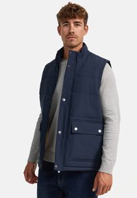 Chaleco acolchado azul marino con un cuello alto, botones a presión plateados y un bolsillo frontal. Combinado con una camisa de manga larga gris claro debajo.