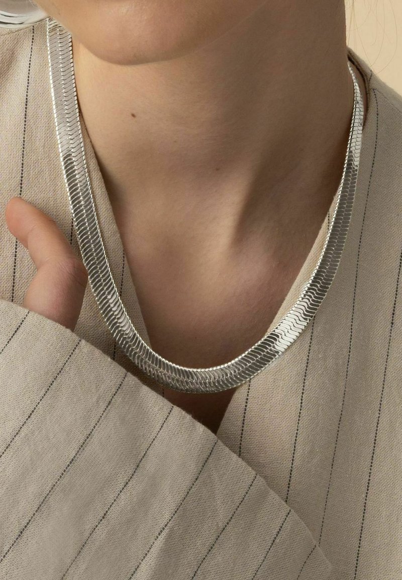 Silverhalsband med en platt, vävd textur, med en slät yta. Bärs mot en ljusrandig tygbakgrund.