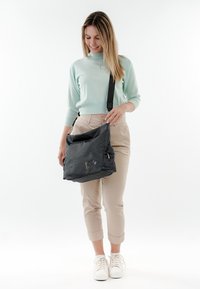 Borsa a tracolla nera in rete con una tasca con zip, cinturino regolabile e dettagli gialli. Il modello indossa un maglione verde chiaro e pantaloni beige.