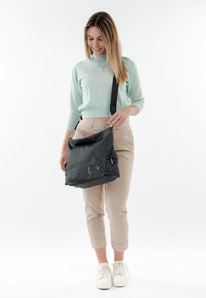 Black mesh crossbody bag with a zippered pocket, adjustable strap, and yellow accents. Model wears a light green sweater and beige pants.