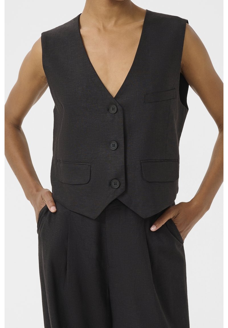 Veste sans manches gris foncé boutonnée de trois boutons, avec deux poches à rabat à l'avant, associée à un pantalon taille haute assorti, mains dans les poches.