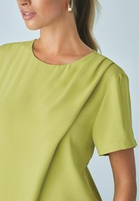 Top lime verde a maniche corte con scollatura rotonda, texture liscia e dettagli di pieghe sottili sulla spalla.