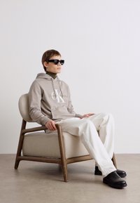 Felpa grigia con logo, pantaloni crema e scarpe nere. Seduto su una sedia chiara con una struttura in legno, di fronte a un muro semplice.