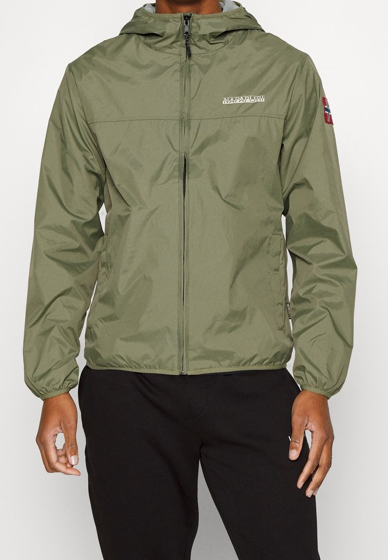 Homme portant une veste à capuche zippée vert olive avec des poignets élastiques et un pantalon noir, debout devant un fond clair uni.