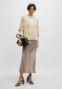 Cardigan lavorato a maglia color crema, caratterizzato da trame in rilievo a treccia e chiusura con bottoni, abbinato a una gonna a sottoveste di seta rosa chiaro e sandali con tacco neri.
