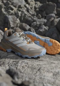 Chaussures de randonnée beiges et marron avec des semelles orange et bleu reposant sur un terrain rocheux, mettant en avant une bonne accroche et un design pour une utilisation extérieure.