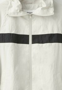 Veste blanche zippée avec une bande noire horizontale sur la poitrine et une capuche avec un cordon.